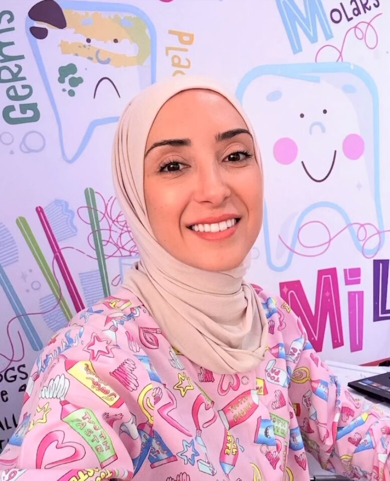 Dr. Basma Monla smiling in front of the clinic's tooth mural